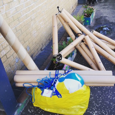 [photo shows a collection of objects on the pavement next to a building. There is a large structure made of cardboard tubes. Several loose tubes of varying sizes and a bright yellow bag with a beach ball poking out of the top}